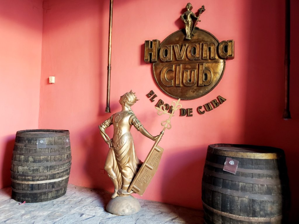 The entrance of Havana Club - Museo del Ron. Two barrels and a bronze statue kept on the floor, with a pink wall forming the background.