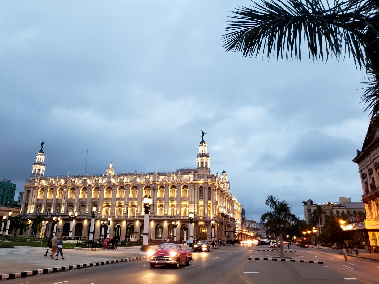 A red colored car with headlights on, a classic building in the background - a street sight of Havana in the evening. Havana is full of such iconic buildings and a must visit in your Cuba 10 days itinerary