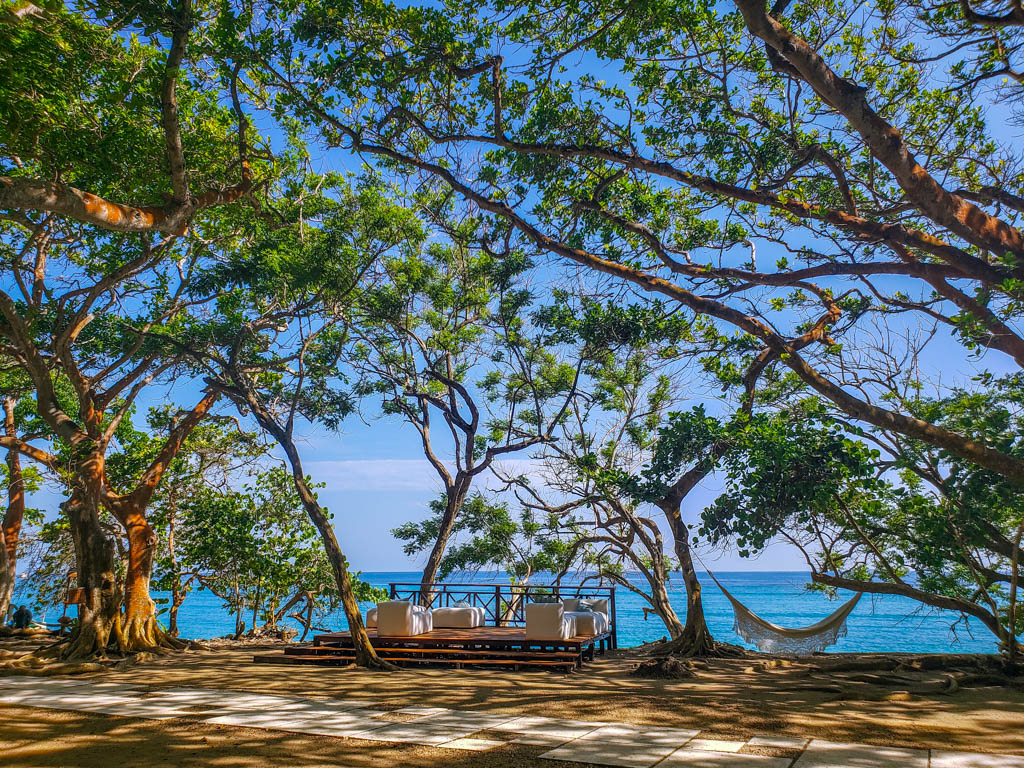 By the blue Caribbean Sea, lies a set of couch on a wooden deck on the hotel property of Hotel San Pedro de Majagua. A hammock is hanging from the tree.
