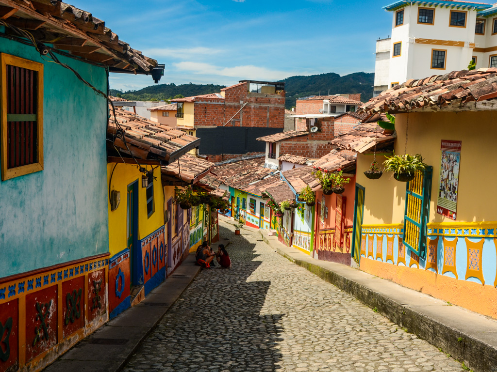 Cobblestone street lined with colorful buildings on either side. The buildings have hanging potted plants and walls with zocalos. 3-4 people sitting on the street, chatting.