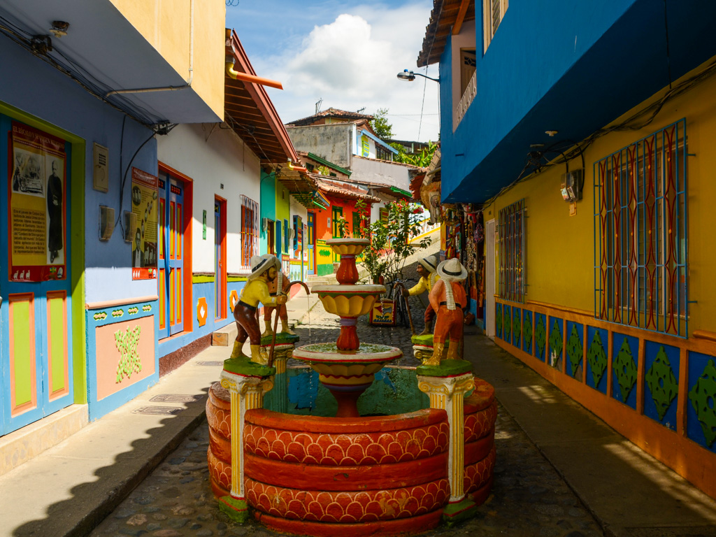 A fountain with sculptures on a street of Guatape. The street is lined with colorful buildings on either side. Guatape day tour.