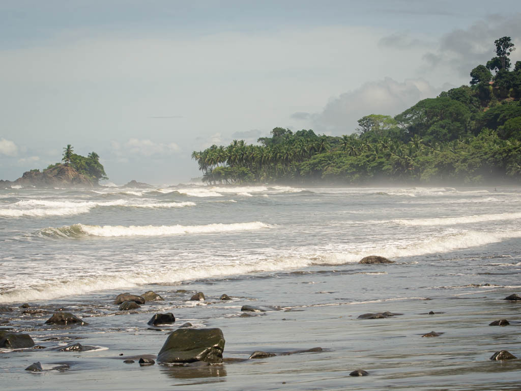 The serene beach with white waves, grey sand and rocks on the beach, lined by lush green vegetation. Playa Dominicalito.