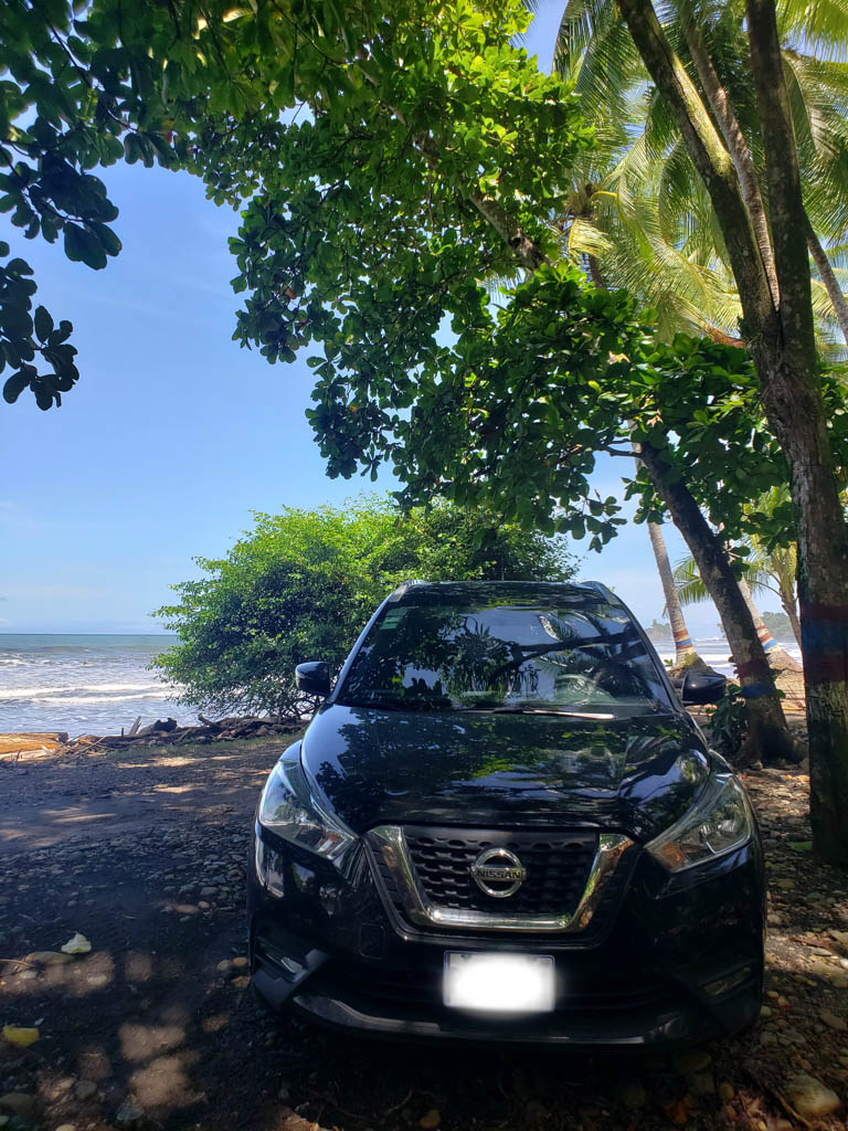 A black car parked near the beach, under the shade of trees.