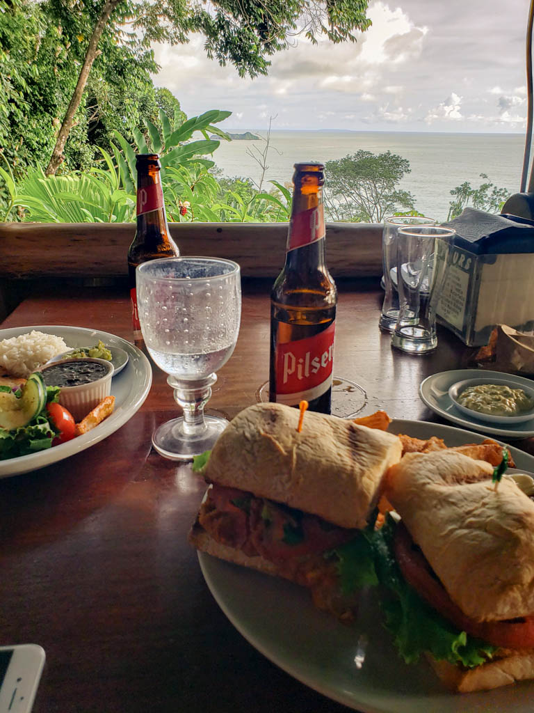 Plates of Casado and Fish Sandwich, two glasses of water two bottles of beer on the table. The window opens to the view of the Pacific Ocean. Lunch at Aracari Restaurant in Uvita.