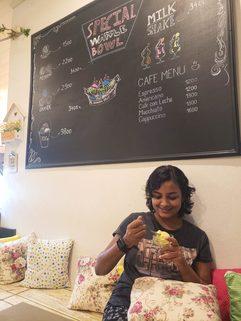 Pubali having a bowl of ice-cream at Gelato Artesanal. The seat is laid out with lots of pillows. Menu on the blackboard on the wall behind.