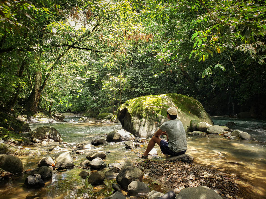 A man, wearing grey t-shirt and blue shorts, sitting on a rock by the river. The river is surrounded by lush jungle.