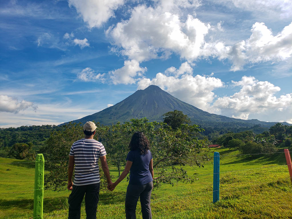 Backside view of a couple holding hands and staring at the Arenal volcano.