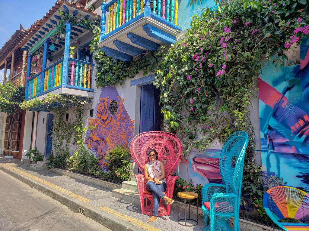 Pubali, sitting on a bright red chair, placed on the pavement next to a wall filled with colorful graffiti. Rainbow colored balcony in the background. Near Trinidad Square.