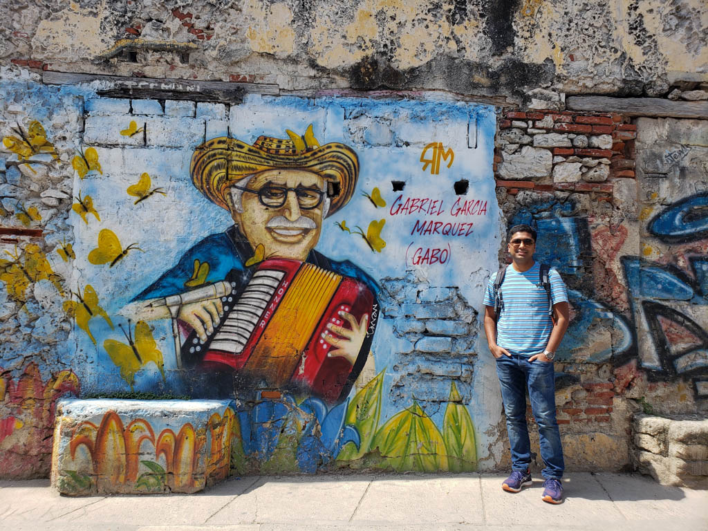 A man wearing blue t-shirt and blue jeans, standing next to a wall depicting Gabriel Garcia Marquez.