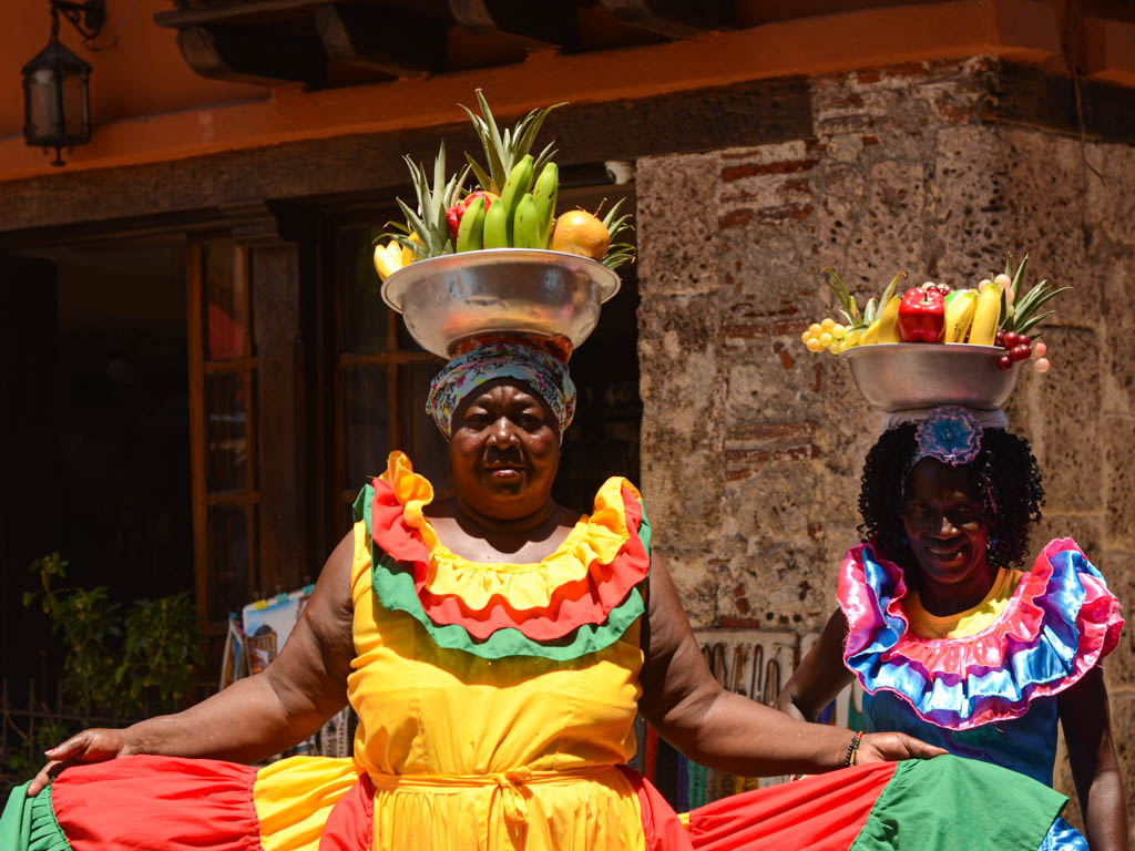 Two Colombian women dressed in colorful traditional dresses, carrying fruits on their heads to sell. They are called Palenqueras.