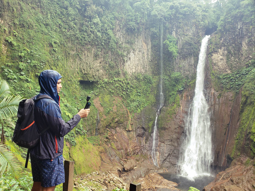 A man wearing blue shorts and blue raincoat, carrying a backpack and holding an action camera, is facing the giant waterfall Catarata del Toro.