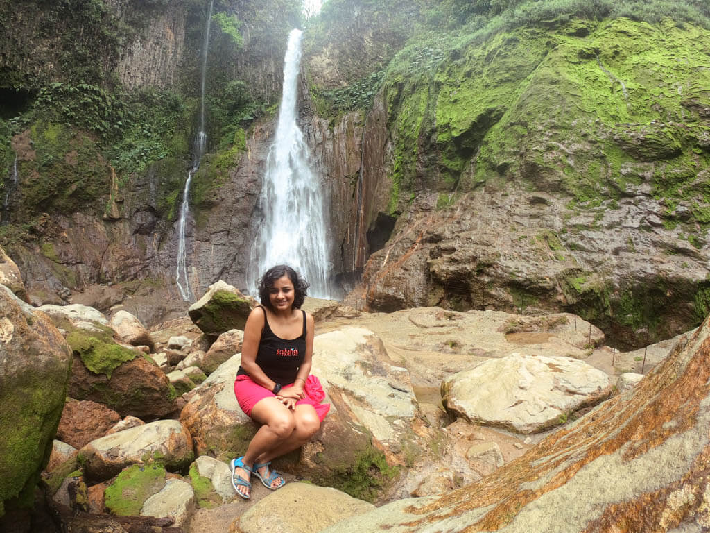 A woman, wearing black top and pink shorts, seated on the rocks next to the waterfall. White waterfall in the background.