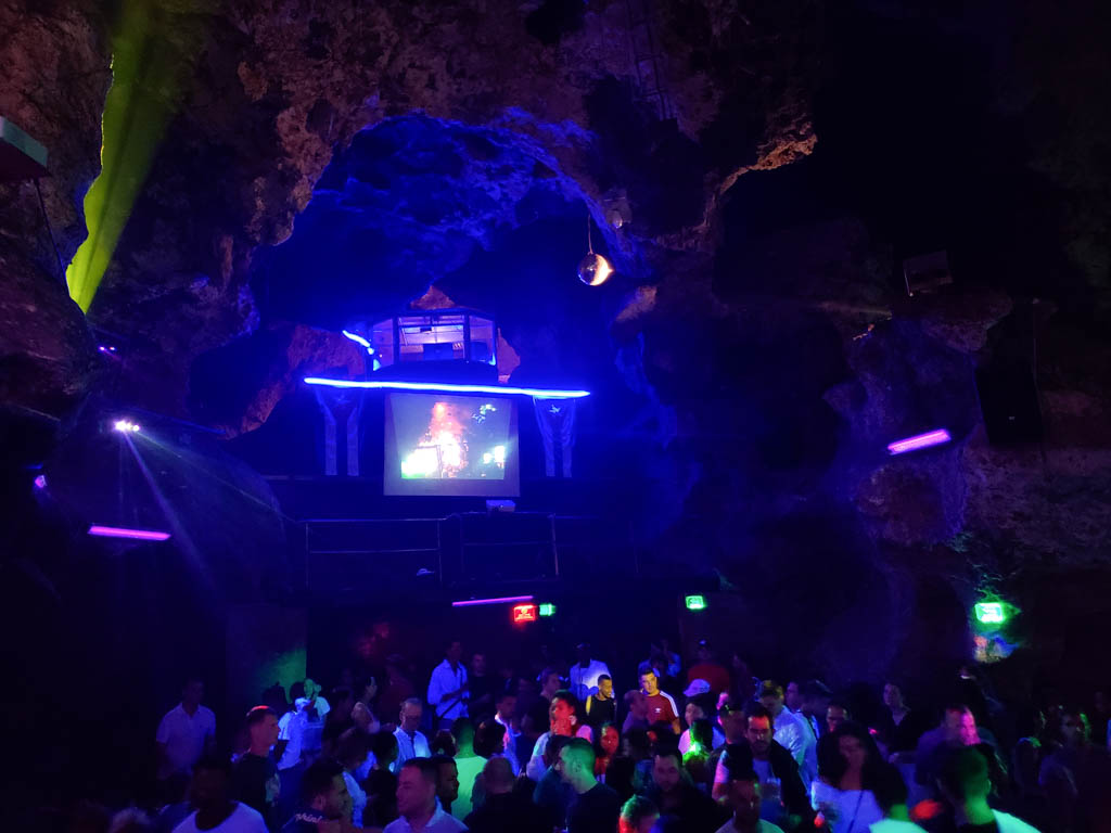 The inside view of Disco Ayala - a cave disco. Dark environment with blue lights. People are dancing on the dance floor.