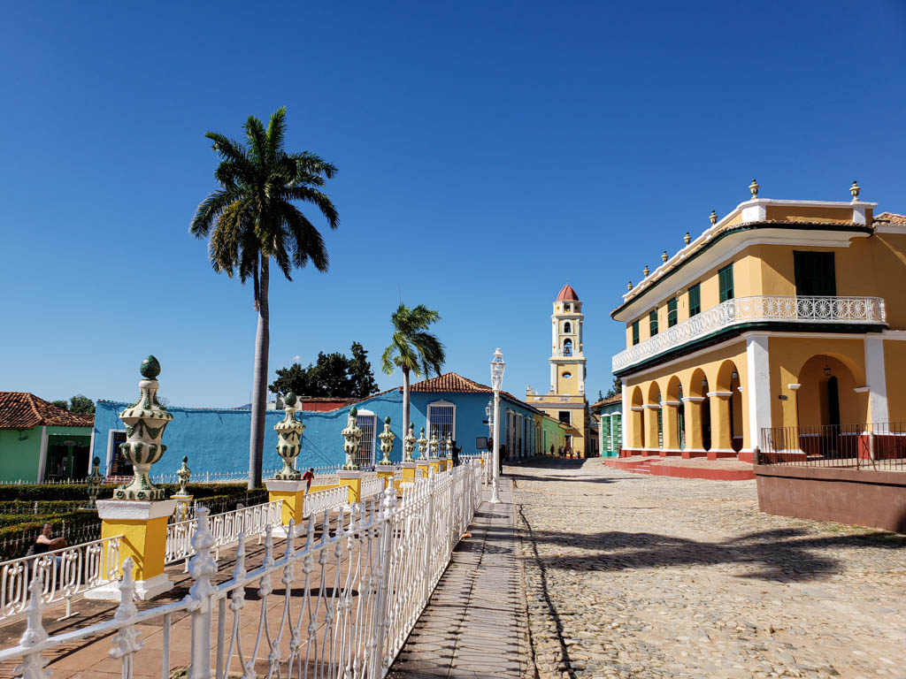 Trinidad, a must visit destination to include on your Cuba 10 days itinerary. Seen in the photo - palm tree, cobblestone street, blue and yellow colored buildings.