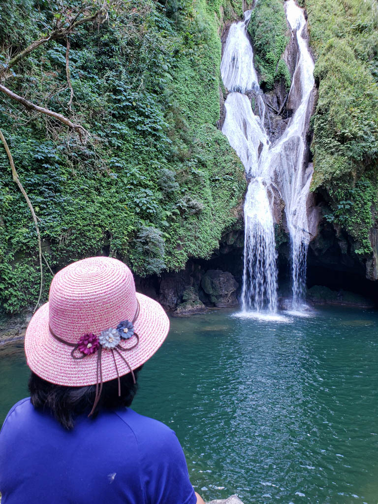 A woman, wearing a pink hat and blue t-shirt, enjoying the view of Vegas Grande Waterfall that is dropping in to an emerald pool.