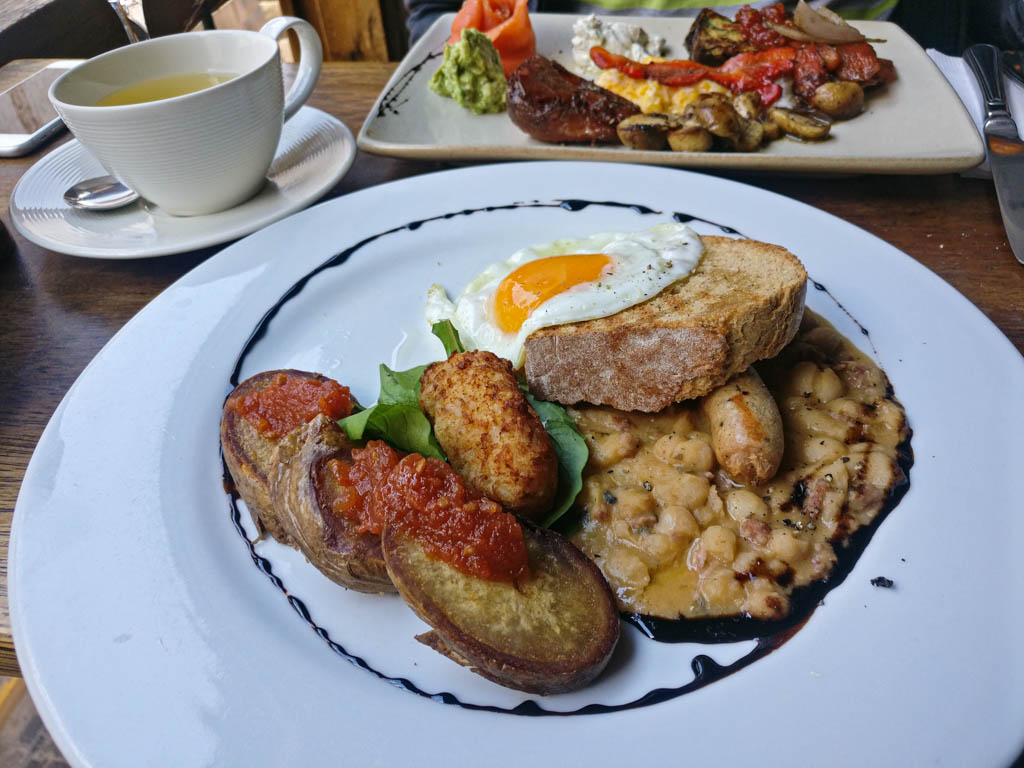 A breakfast plate with eggs, bread, beans, sausages, accompanied with a cup of cocoa tea.