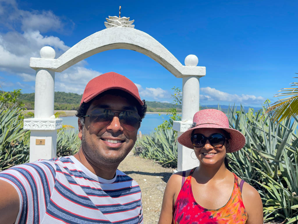 Man wearing red striped white t-shirt, red baseball cap and sunglasses, woman wearing tie dye spaghetti top, pink hat and sunglasses, posing at Cabuya Island Cemetery entrance, with the ocean in the background.