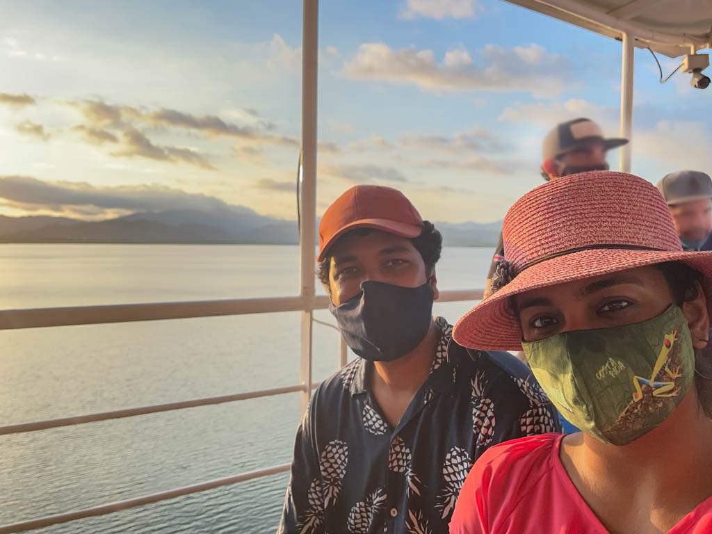 Man wearing red cap, dark blue floral printed shirt and dark blue mask, and woman wearing pink dress, pink hat and green mask, seated on the upper deck of Puntarenas Ferry. Seen in the background, the sky and the gulf waters.