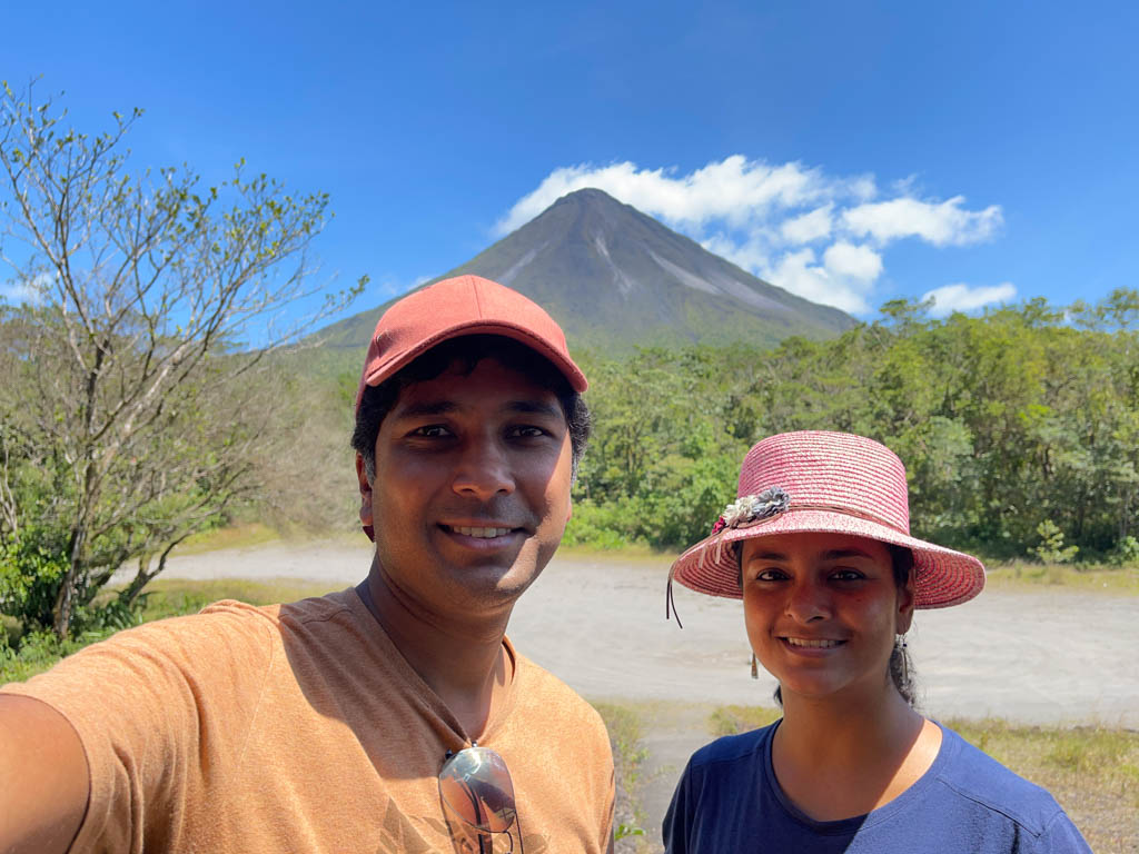 Man wearing beige t-shirt and red cap, and woman wearing blue t-shirt and pink hat, posing for a selfie with Arenal Volcano in the background.