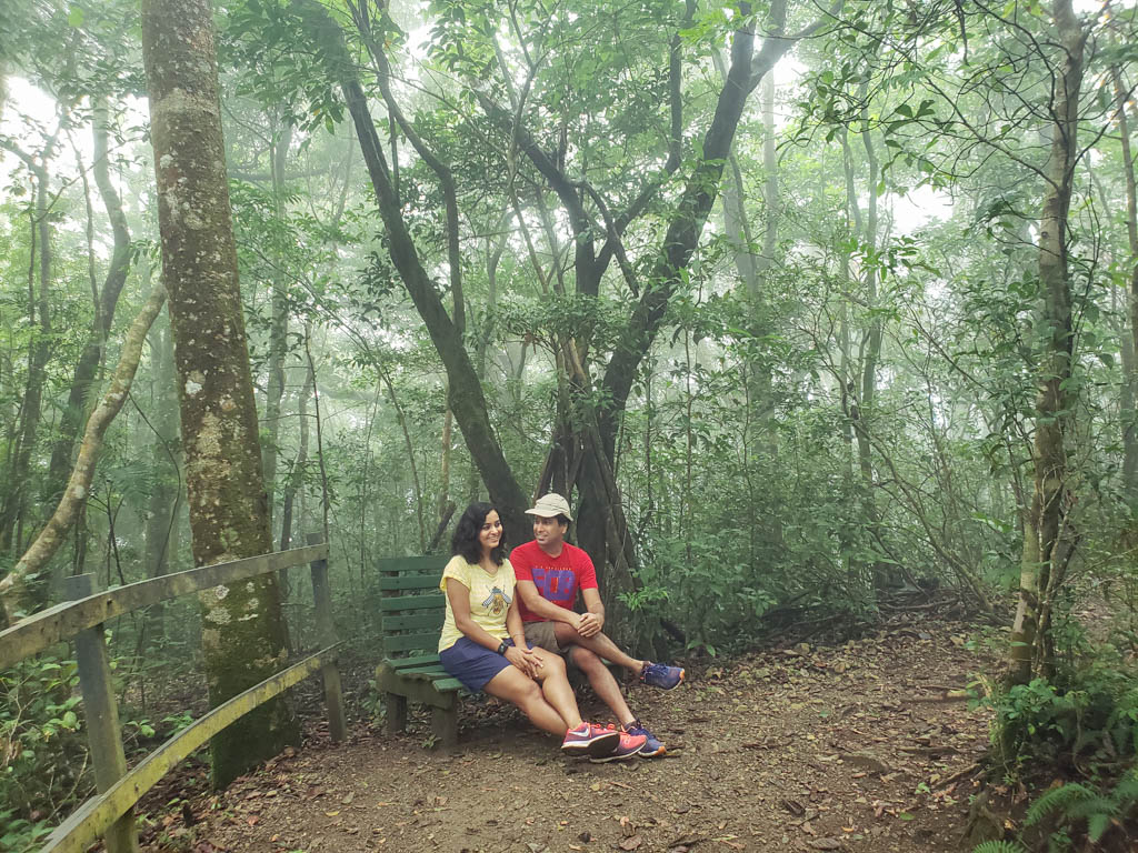 Woman wearing yellow t-shirt, navy blue short skirt and peach sneakers, and man wearing red t-shirt, hiking shorts and blue sneakers, sitting on a bench in the dense cloudforests of Monteverde, Costa Rica.