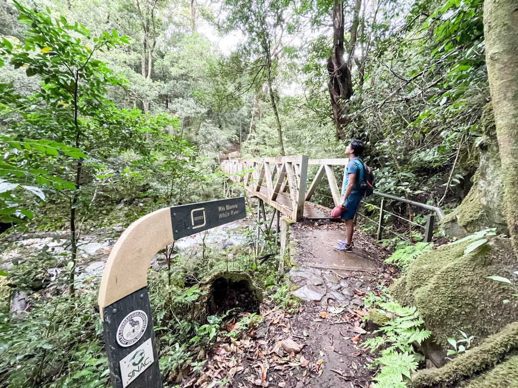 A man wearing blue shorts and blue t-shirt, and carrying a small backpack, is standing near the river bridge over Rio Blanco, that is part of La Cangreja Waterfall trail.