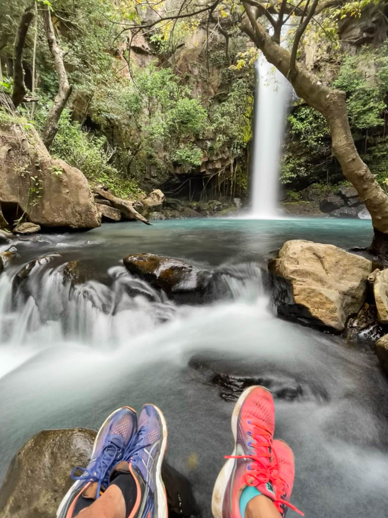 Two pairs of feet, wearing blue and red sneakers respectively, with La Cangreja Waterfall and the the blue pool in the background.