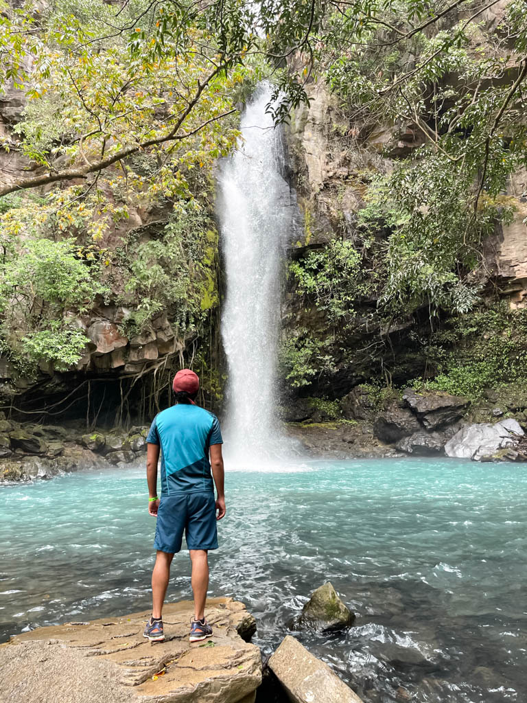 A man wearing blue shorts, blue dry-fit t-shirt, red baseball cap and blue sneakers, looking at La Cangreja Waterfall, appreciating the view.
