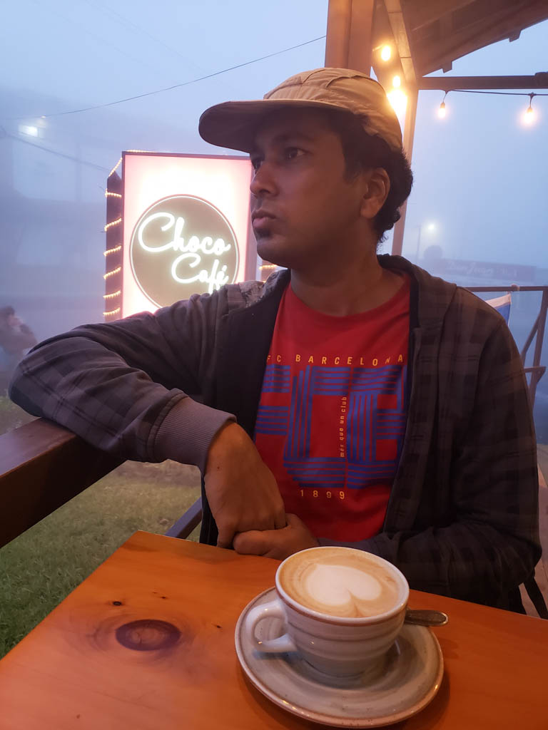 Man wearing red t-shirt, dark grey jacket, beige cap, looking out of a coffee shop in a thoughtful mood. A cup of cappuccino in front on the table.