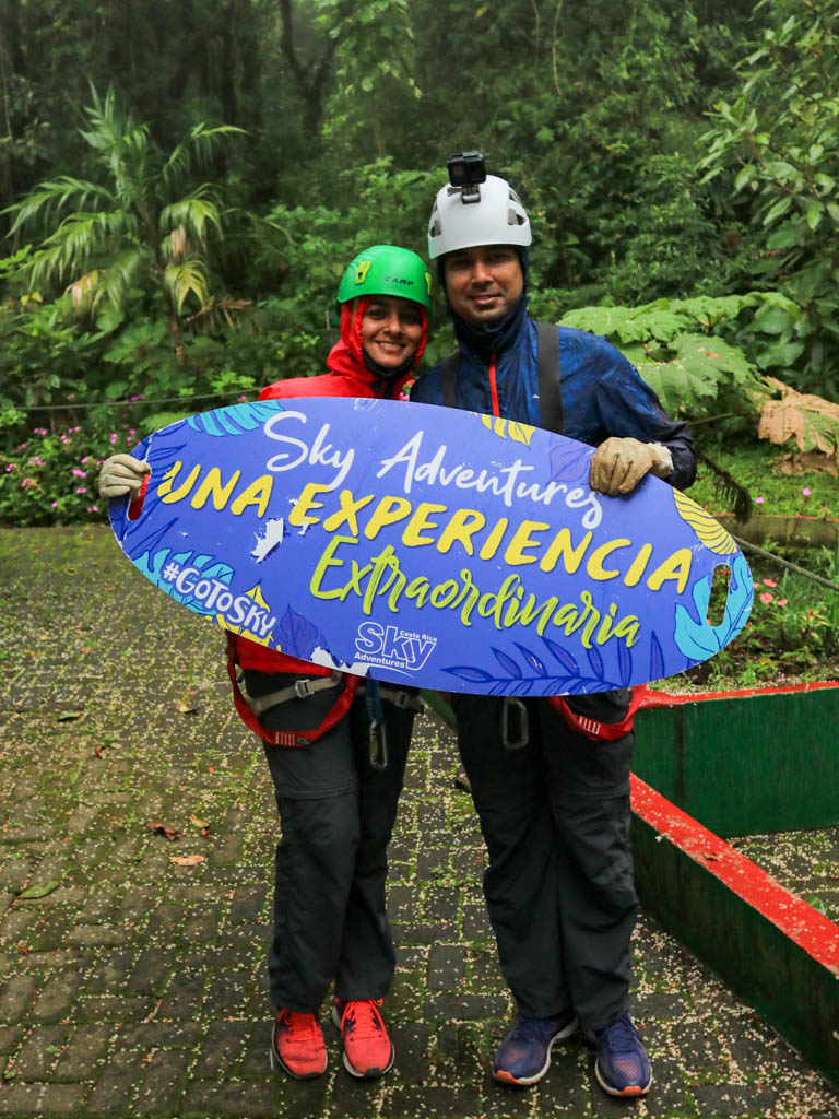 A man and woman geared up for zip lining and posing with a placard of Sky Adventures as a memento.