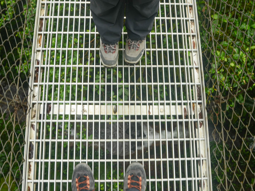 Feet of a man and a woman on a hanging bridge with a stream flowing below.