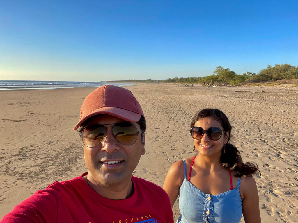 A man wearing red t-shirt, sunglasses and red cap, and a woman wearing a denim dress and sunglasses, posing for a selfie at Playa Avellanas in Guanacaste, Costa Rica.