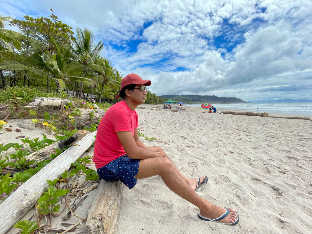 A man sitting on a tree log at a white sand beach framed by blue sky. The man is wearing red cap, red t-shirt, blue swim shorts and sunglasses.