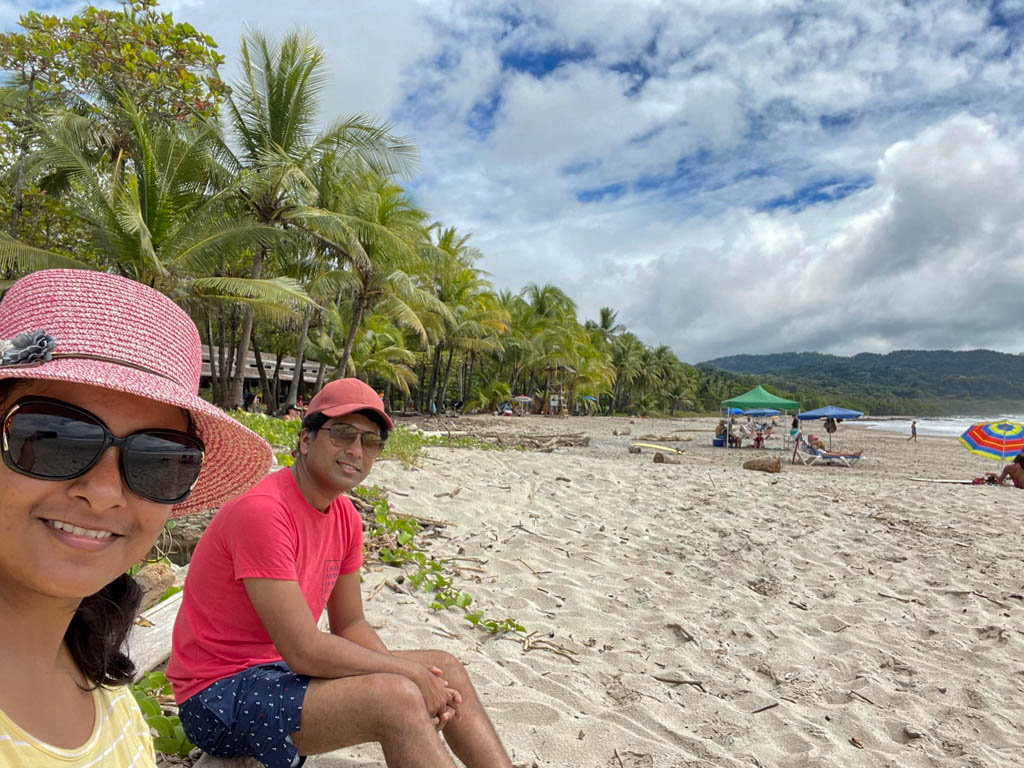 A couple posing for a selfie at a alm-fringed white sand beach framed by blue sky at Playa Carmen, Costa Rica. The man is wearing red cap, red t-shirt, blue swim shorts and sunglasses. The woman is wearing yellow top, pink hat and sunglasses.