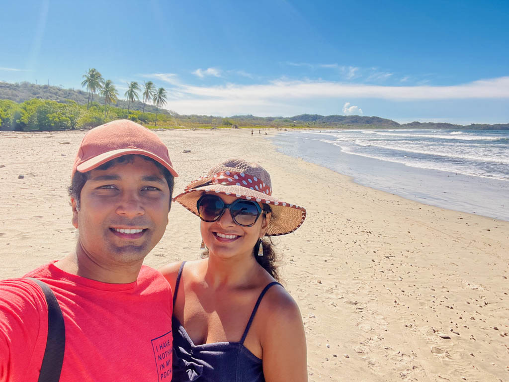 Man wearing red t-shirt, red cap and woman wearing beach hat, sunglasses and denim spaghetti top, posing for a selfie at Playa Guiones. A vast stretch of the beach in the background.