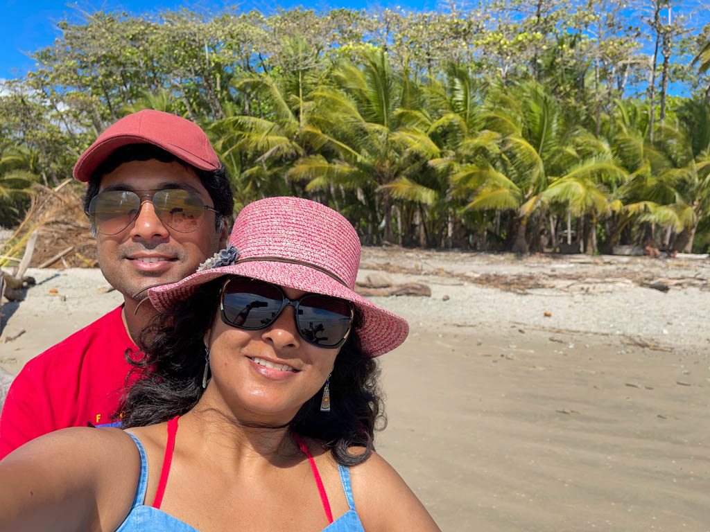 Man wearing red t-shirt, red cap and sunglasses, and woman wearing pink hat, blue dress and sunglasses, posing for a selfie at the beach with palm trees in the background.