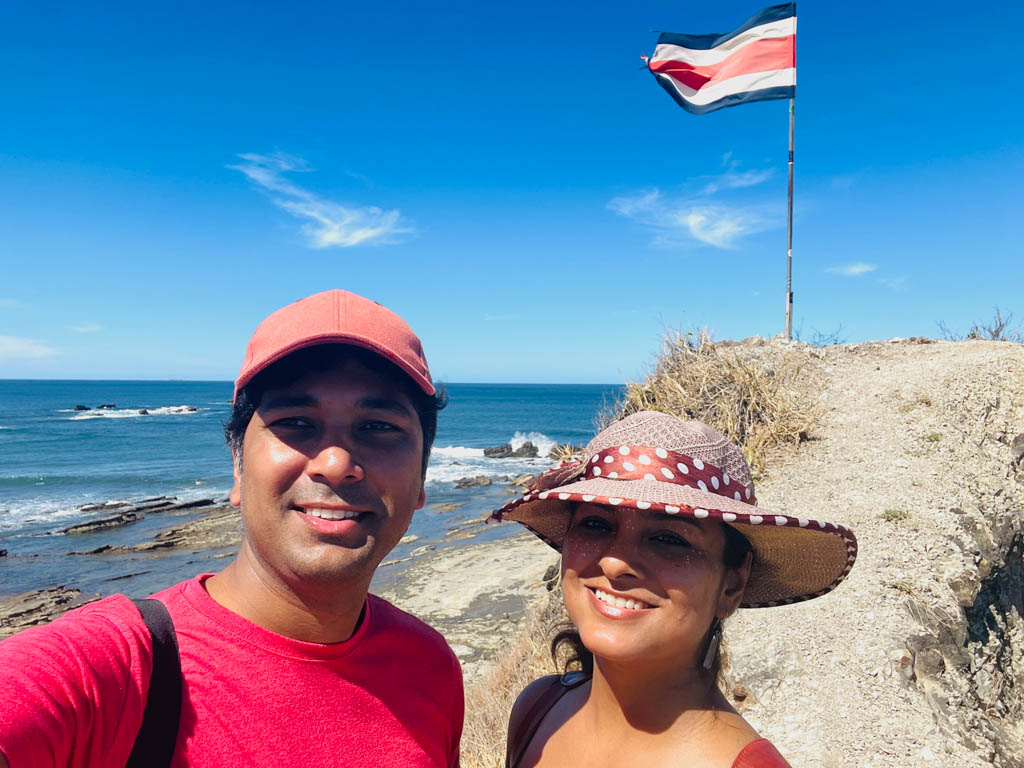 Man wearing red t-shirt, blue shorts and red cap, and a woman wearing a beach hat, posing for a selfie on the ridge of a cliff. Costa Rica flag pole and Pacific Ocean view in the background.