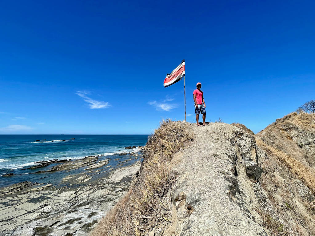 Man wearing red t-shirt and blue shorts standing on the ridge of a cliff. Costa Rica flag pole next to the man. Pacific Ocean view in the background.