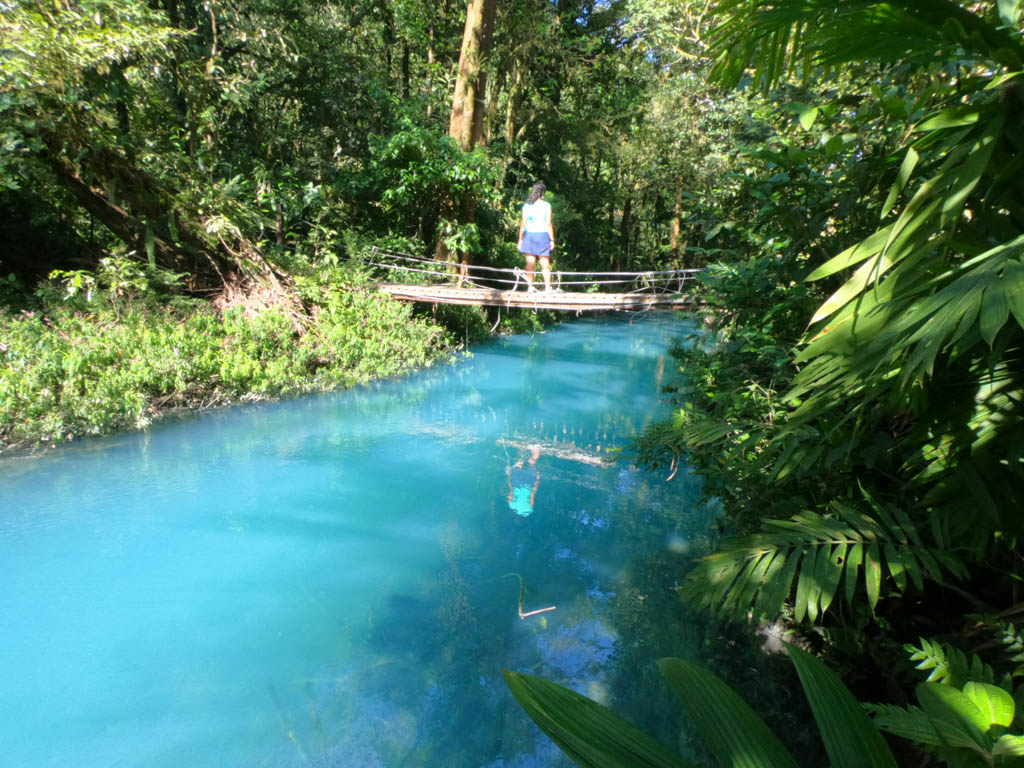 Woman wearing sark blue skirt and green dry-fit tee, standing on the wooden hanging bridge over the blue Rio Celeste.