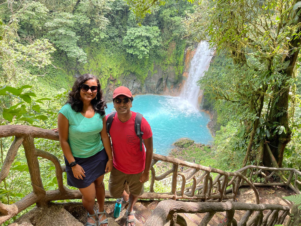 Paradise Catchers posing on the staircase leading to the Rio Celeste waterfall, with the waterfall in the background. Man is wearing red cap, sunglasses, red t-shirt and hiking sandals. Woman is wearing sunglasses, green dry-fit tee, dark blue skirt, fitness watch and hiking sandals.