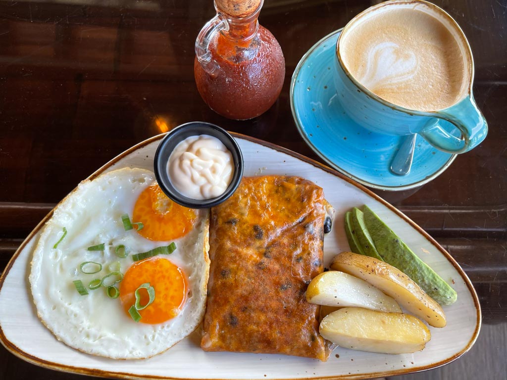 A breakfast plate containing cheese roll wrapped gallo pinto, sunny side up, potatoes and avocadoes. A blue cup of coffee on the side. At Fuego restaurant in Dominical.