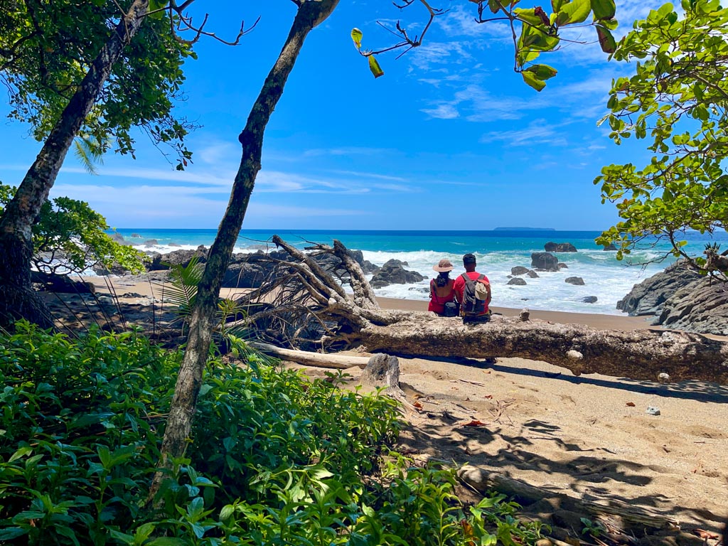 A man and a woman, seated on a fallen tree log, along the Drake Bay Beach Hike Trail, enjoying the view of the Pacific Ocean and the Caño Island in the distant horizon.