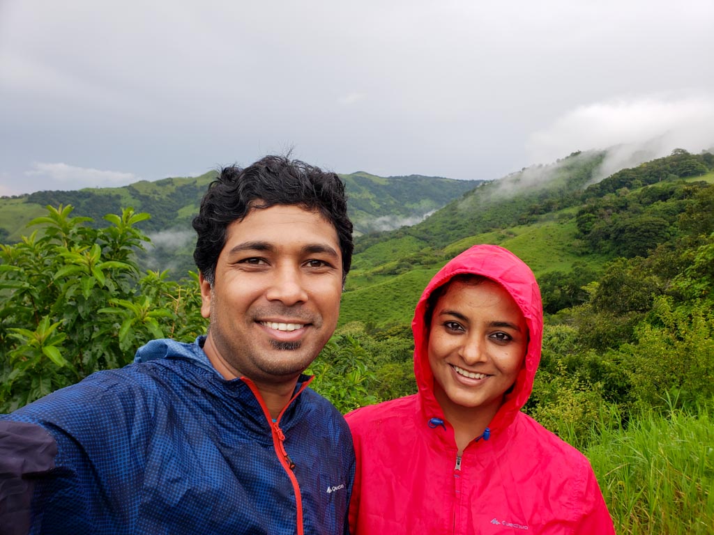 A man and a woman posing for a selfie with a backdrop of mountains with lush vegetation. Rainy season in Monteverde.