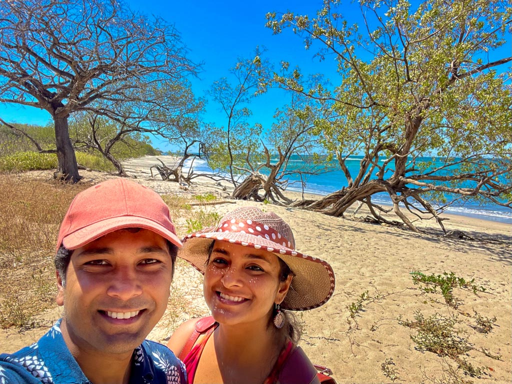 A man wearing blue and white floral shirt and red cap, and a woman wearing a red spaghetti top and brown hat, posing for a selfie at Playa Lagartillo in Guanacaste, Costa Rica.