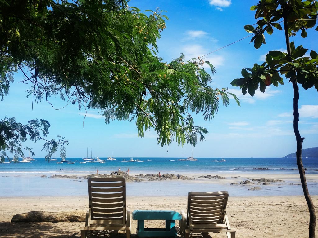 Two empty beach chairs under the shade of trees. They are facing the blue ocean, where many yachts are anchored. This is a typical sight at Tamarindo on quieter mornings. Tamarindo is a popular beach destination on Costa Rica itineraries.