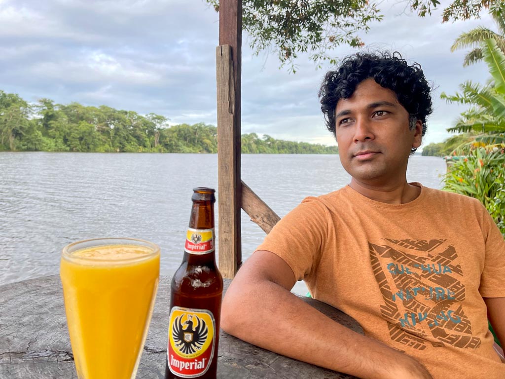 Man seated at a waterfront table that has a glass of mango juice and a bottle of beer.