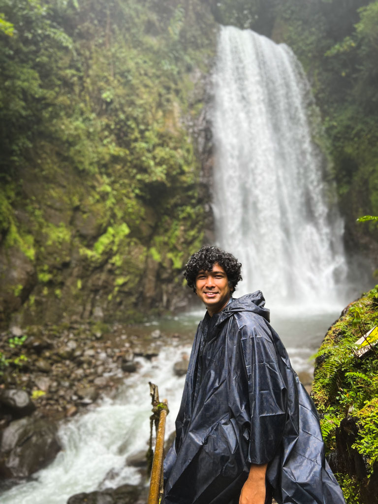 Man wearing a rain poncho, standing near the waterfall. Rain Poncho is a packing essential for Costa Rica in the rainy season.