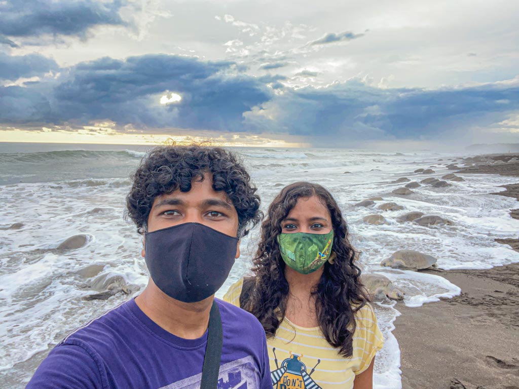 A man and a woman posing for a selfie at Playa Ostional, with several olive ridley sea turtles in the background.