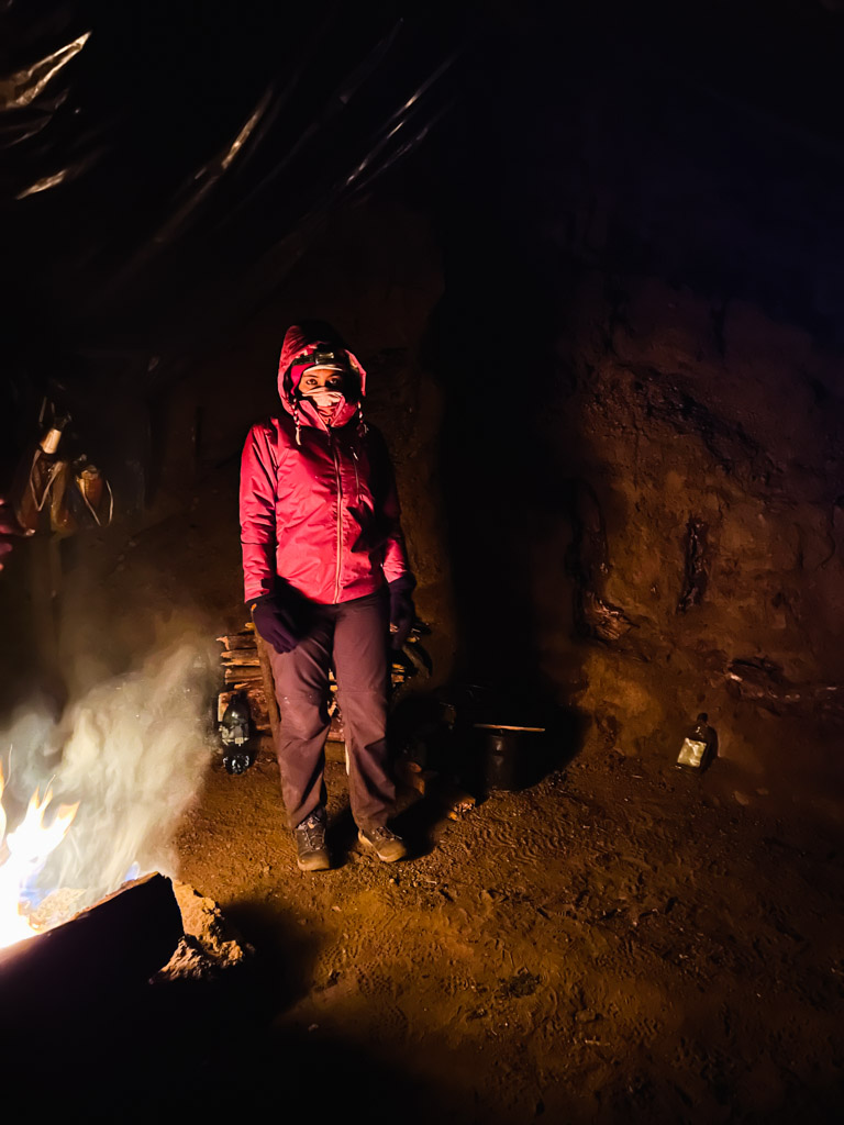 Woman wearing hiking boots, hiking pants, jackets, caps, hood, bandana and gloves, standing next to the campfire at Acatenango base camp. All these items are essential while packing for Acatenango hike.