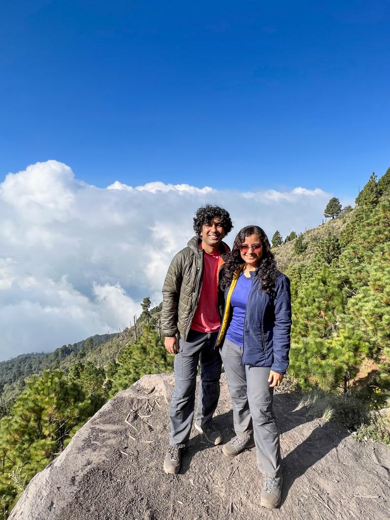 Man ans woman wearing hiking pants, quick-dry t-shirts, hiking boots, jackets and sunglasses, standing at La Pena, a protruded rock on the Acatenango hiking trail.