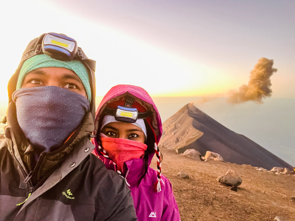 At Acatenango volcano summit, man and woman, wearing multiple layers of clothes, bandanas and hoods, and flashlights, all essential items when packing for Acatenango hike. Fuego volcano in the background.